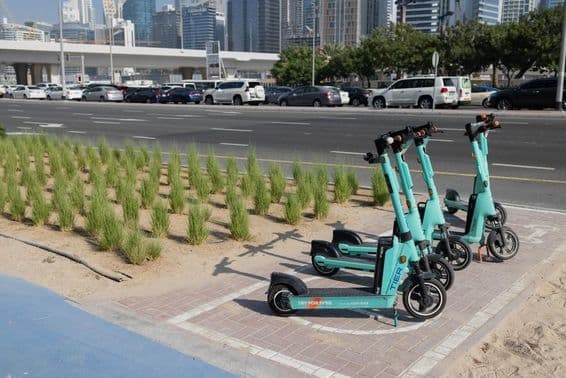 E-scooters parked in front of Dubai skyscraper with green plants on road divider.