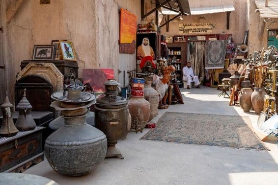 Market scene with antique vases, carpets, pictures, and a seller seated in the background waiting for customers.