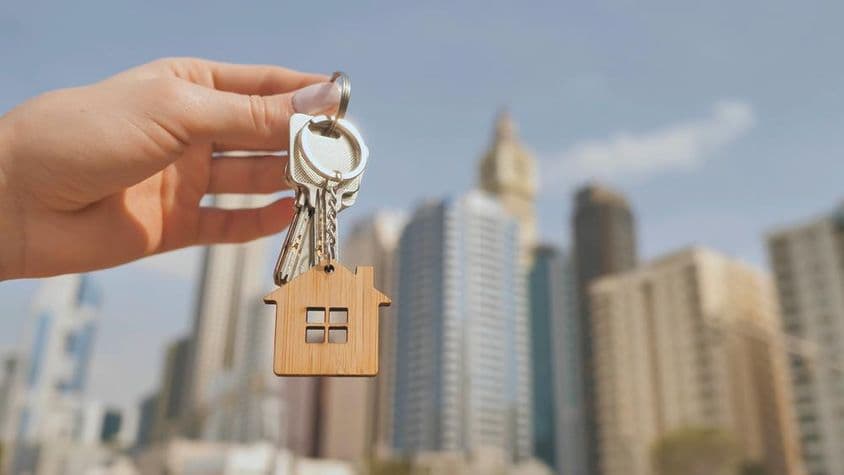 A key with a wooden house keychain, blurred Dubai skyscrapers in the background.
