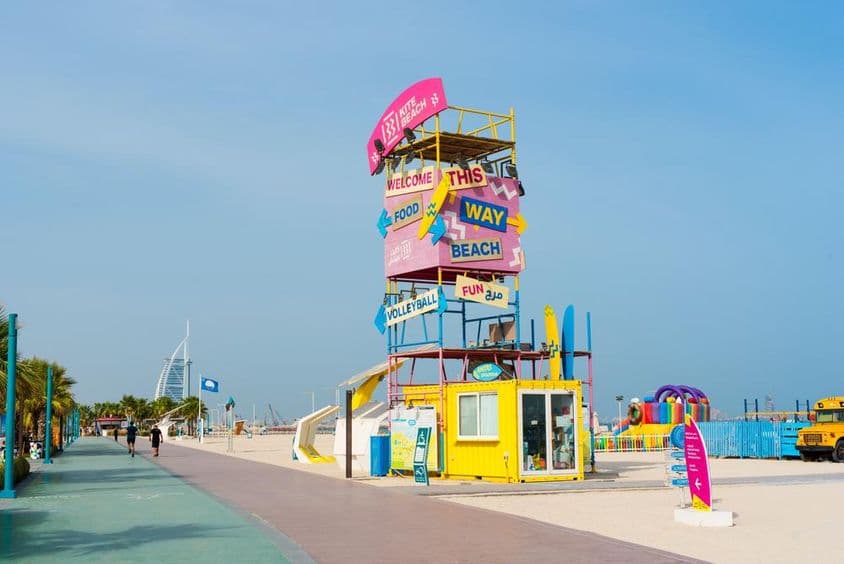 Dubai Kite Beach welcome structure, signs, Burj Al Arab in background, foreground with running track and sandy beach.
