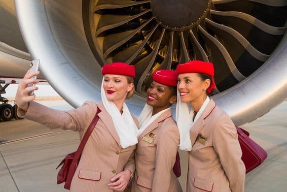 Three Emirates flight attendants taking a selfie in front of an Emirates plane engine.