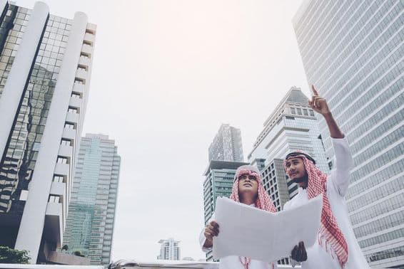 Two Arab designers with a blueprint gaze into the distance, skyscrapers behind them.