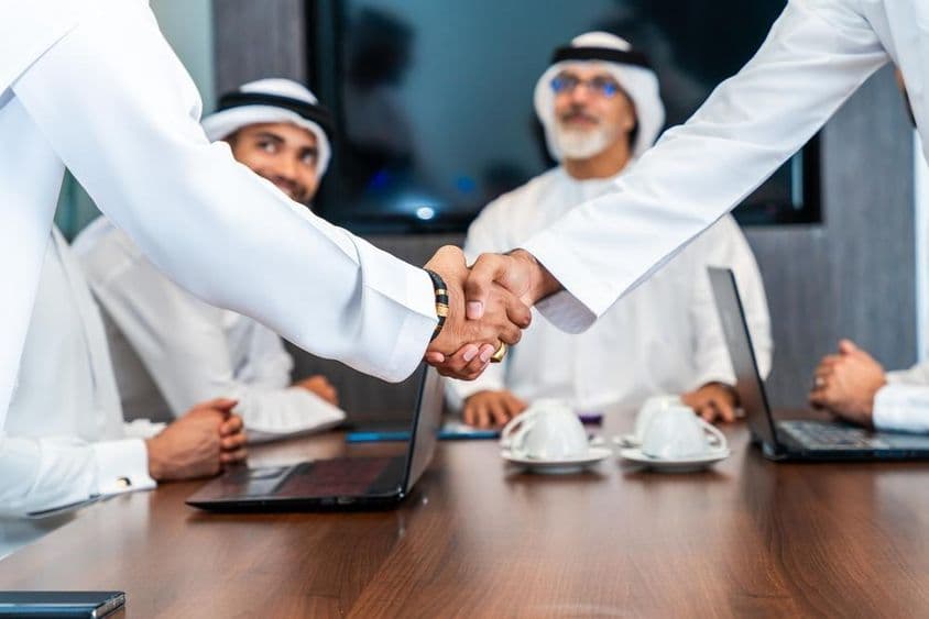 Emirates executives shake hands at a conference table, with phones, notebooks, and coffee cups on the table. Two Emiratis are smiling in the background.