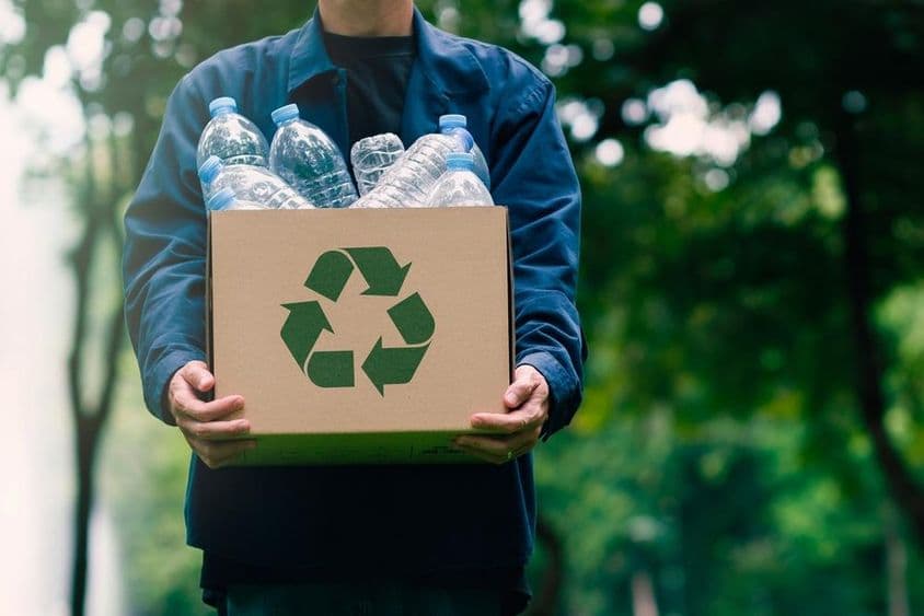 A person carrying a cardboard box with returnable bottles, with blurred green trees in the background.