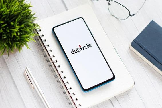 A phone running Dubizzle on a table, under a notepad, next to glasses and a small green plant.