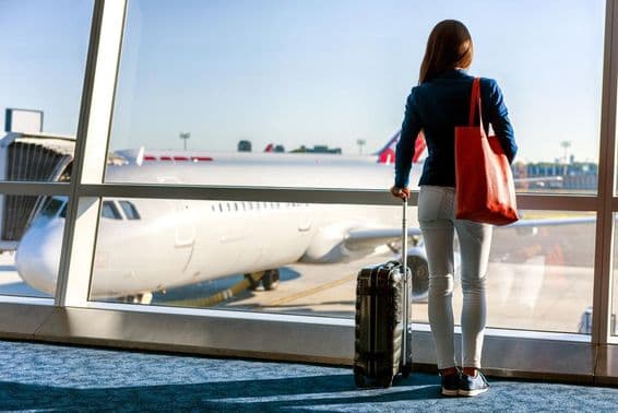 A woman stands by the window with a visible airplane, holding a rolling suitcase with her left hand and a handbag in her right.