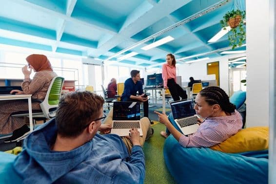 People working in an office, two individuals sitting on beanbags with laptops on their laps.