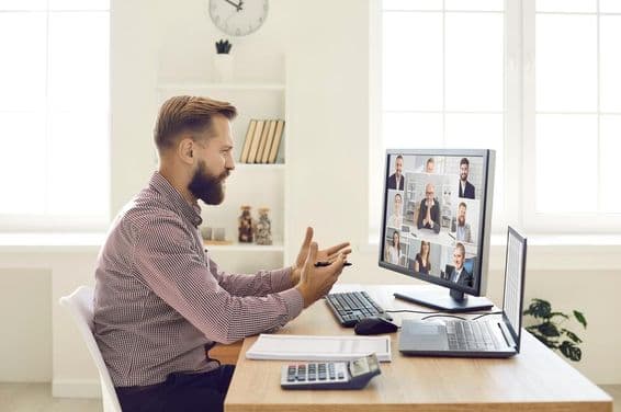 A person in an office setting with a notebook and calculator on the desk, conducting a video conference on a monitor with a webcam.