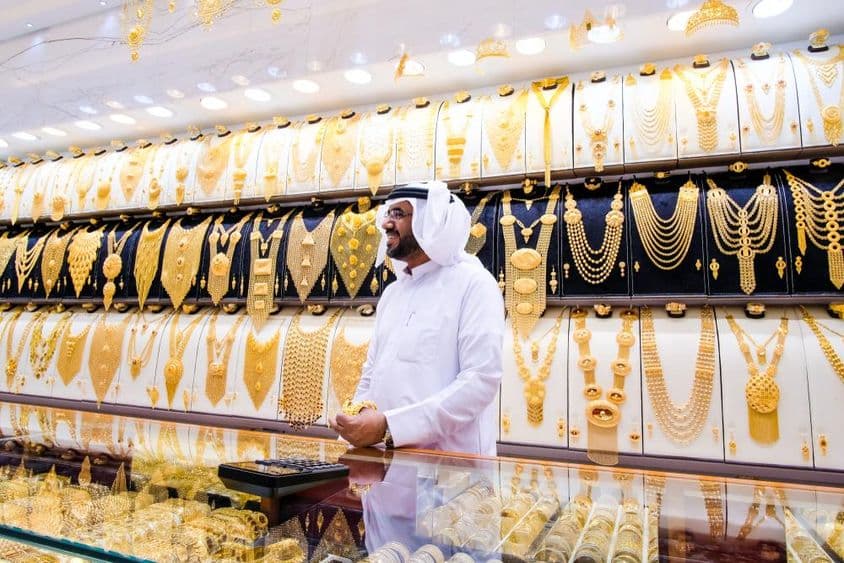 Dubai gold vendor, with a background of many gold necklaces, the vendor in traditional white attire.
