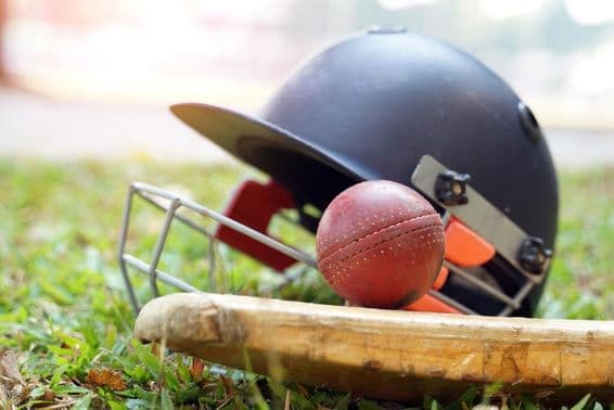 A cricket bat with a ball above and behind it, a cricket helmet is visible on a grassy field.