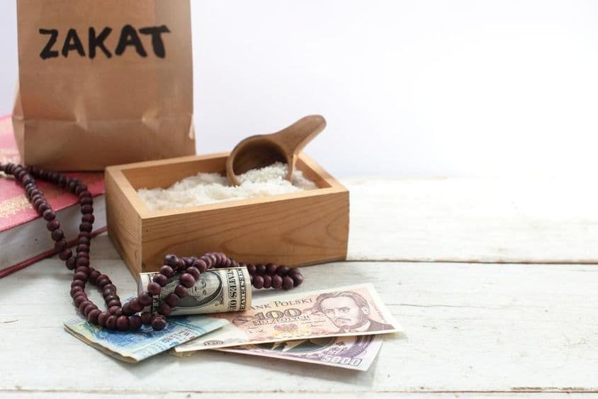 Zakat labeled bag in front of a box with rice, pearl beads, dollars, and other currencies.