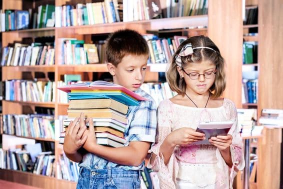 Two children in the library, one reading an e-book, the other carrying many books in their hands.