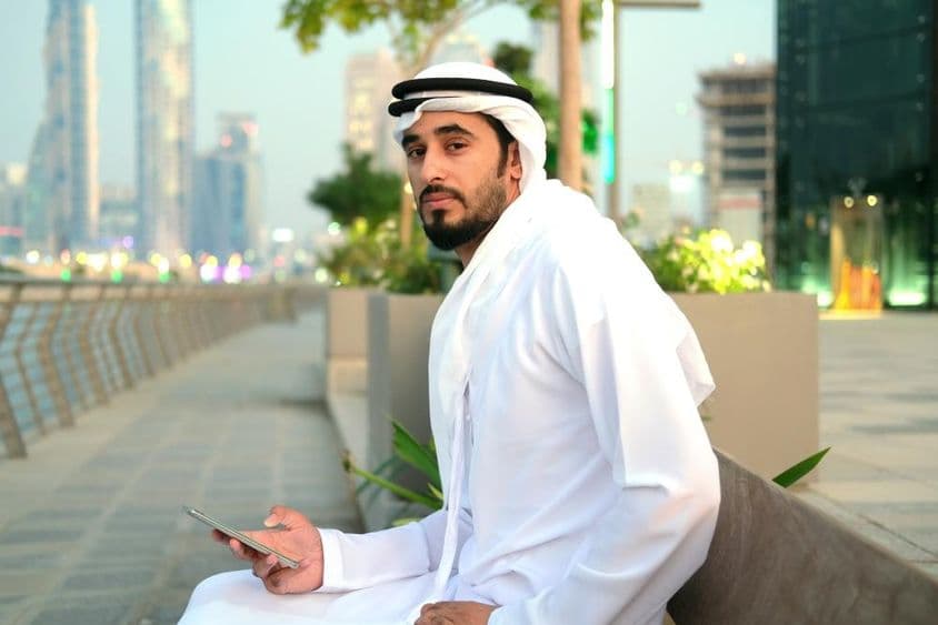 Emirati citizen sitting on a bench, holding a phone, skyscrapers in the background.