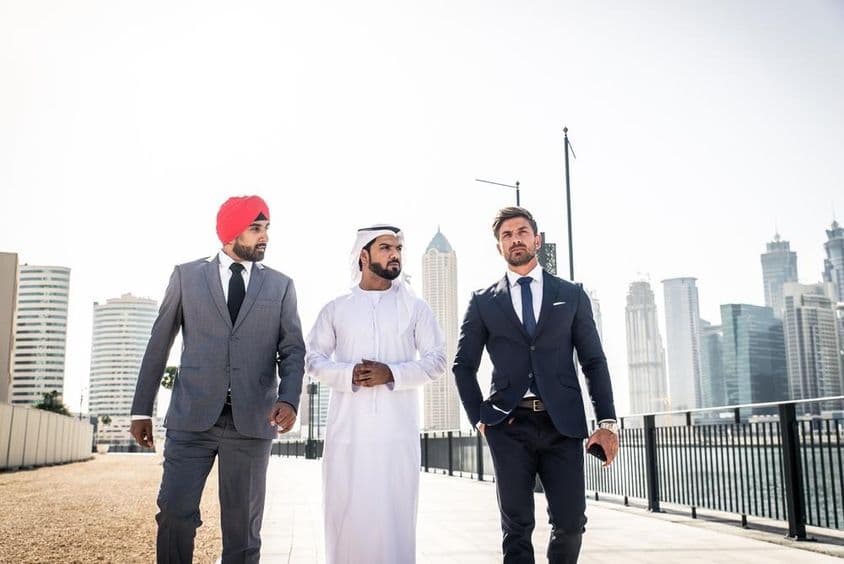 An Indian, an Emirati, and a European citizen walking along the canal, skyscrapers behind them.