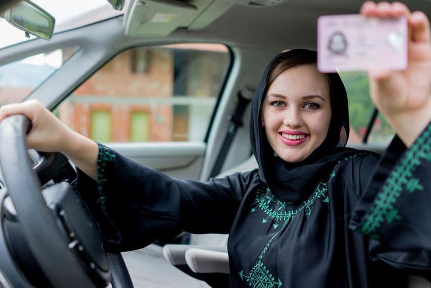 A lady in Dubai holds a driver's license with one hand and the steering wheel with the other, smiling.