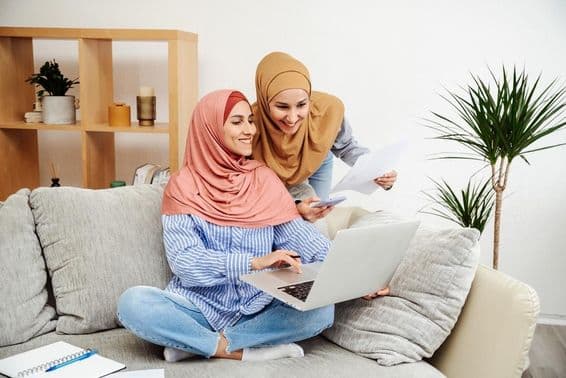 Two Emirati women reviewing a contract and notebook, one holding a phone, sitting on a couch.