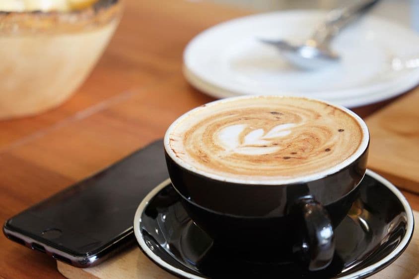 A cup of cappuccino on a table with a phone beside it.