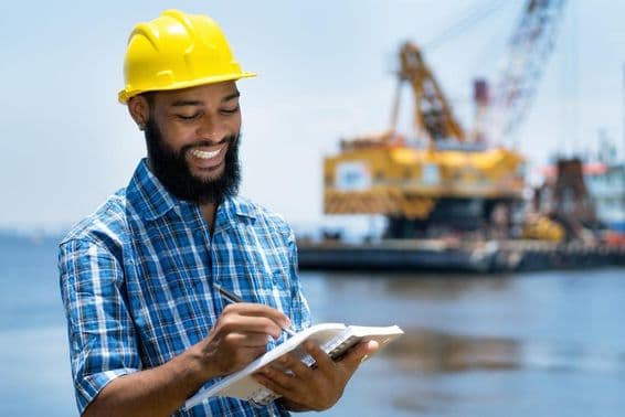 Nigerian male worker jotting notes on the beach, a crane being carried by a transport vehicle sleepily in the background.