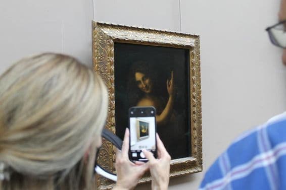 The painting of St. John the Baptist on a wall, with a woman taking a photo of it with a phone.