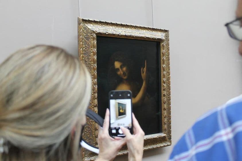 The painting of St. John the Baptist on a wall, with a woman taking a photo of it with a phone.