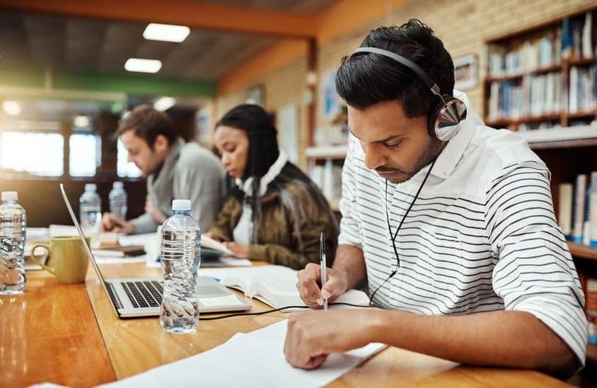 College students writing on paper with book and notebook in front, one wearing headphones.