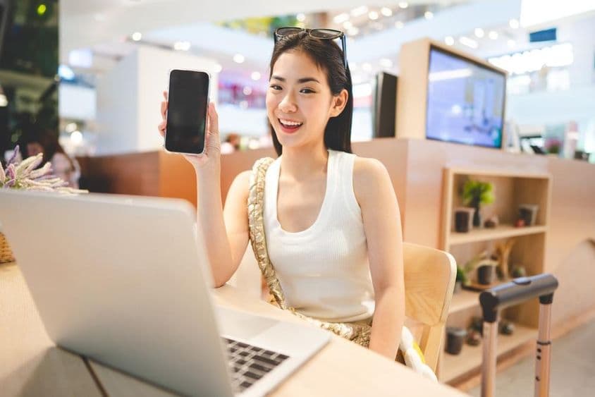 Gen Z girl working, holding a phone, with a notebook in front of her and a rolling suitcase handle beside her.