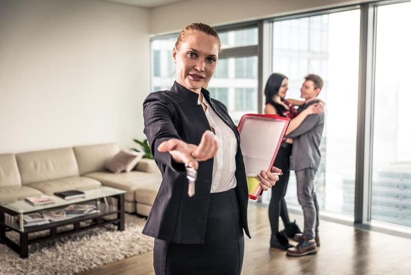 A female real estate broker waving, a couple hugging in the background.