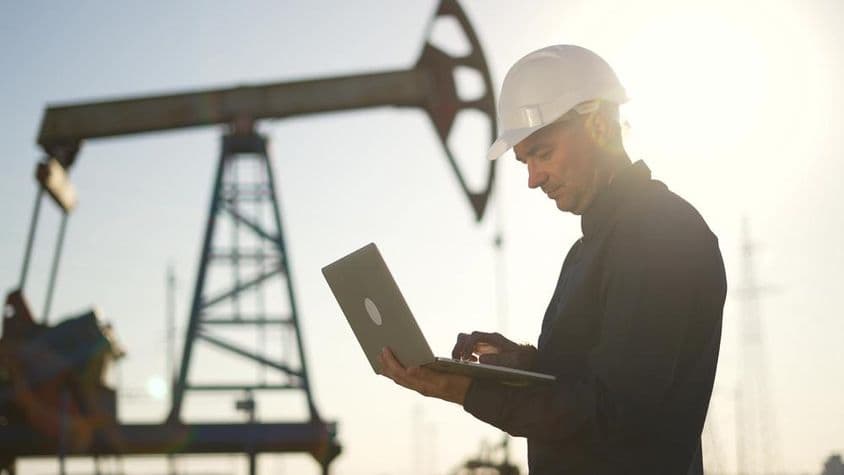 An oil drilling rig in the background with a worker using a notebook in the foreground.