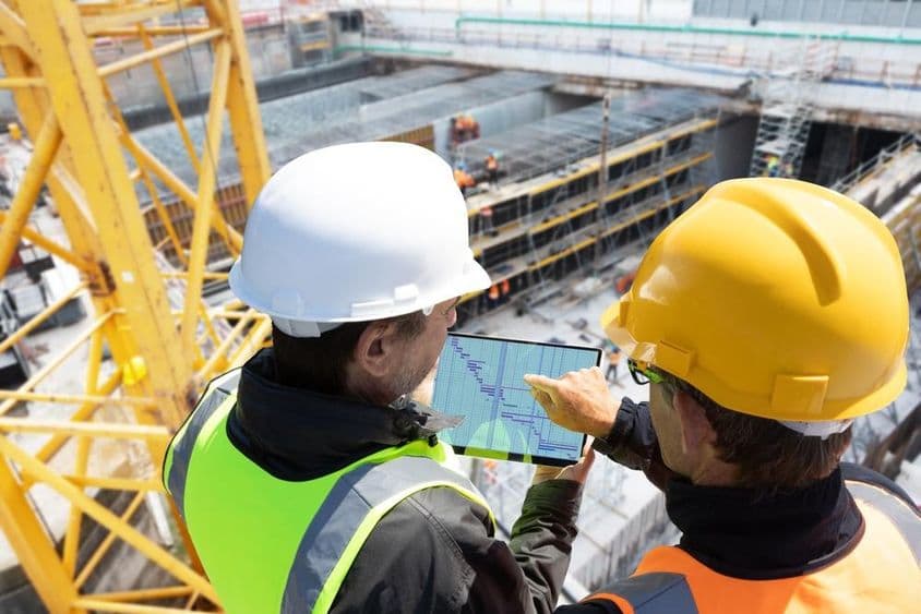 Two people in helmets standing on a crane at a construction site, checking plans on a tablet, with the construction site in front of them.