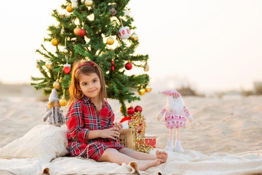 Happy smiling little girl sitting on the beach under a Christmas tree.
