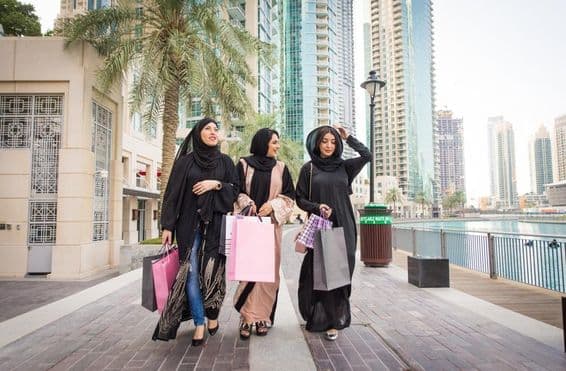 3 Emirati women walking with shopping bags after shopping, with skyscrapers in the background.