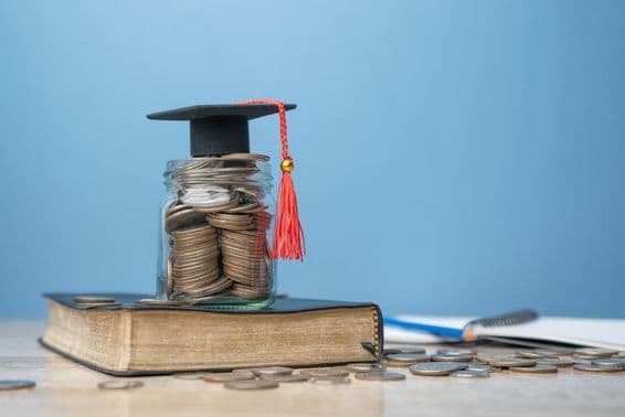A tiny graduation cap atop a jar filled with coins, placed on a book.