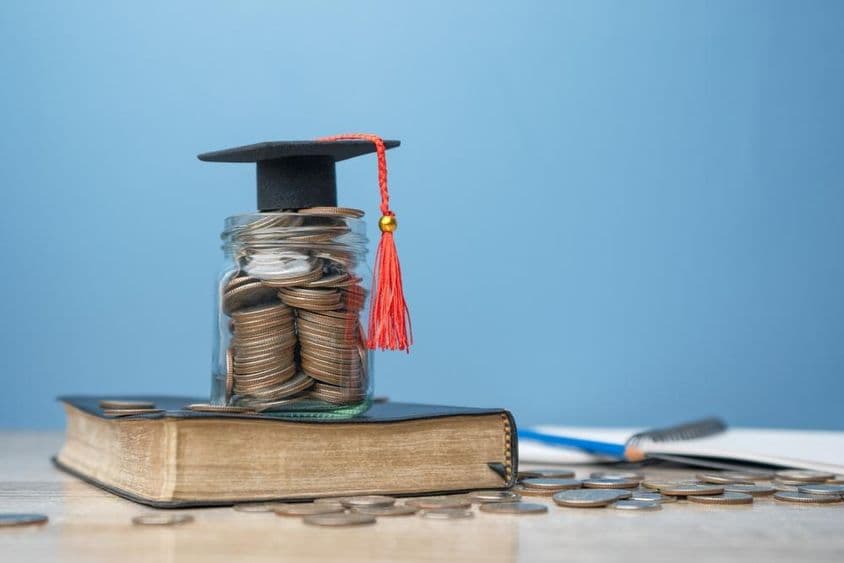 A tiny graduation cap atop a jar filled with coins, placed on a book.