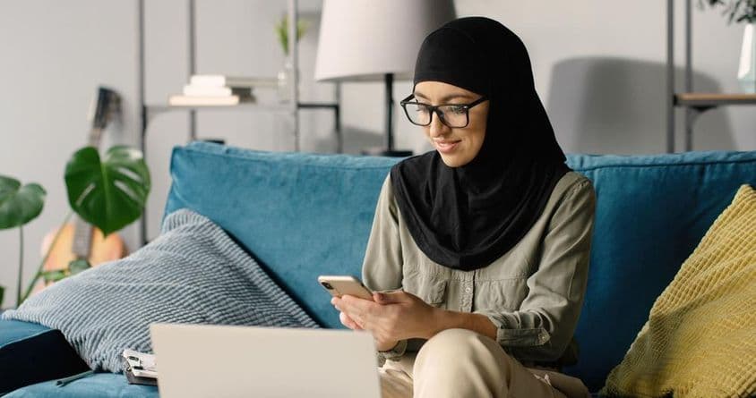 Arab girl in hijab working at home using smartphone and laptop while sitting on the couch.