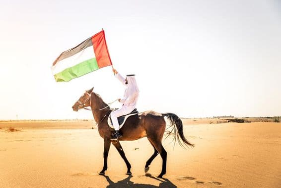 Middle Eastern man in traditional Emirati clothing riding an Arabian horse in the Dubai desert.