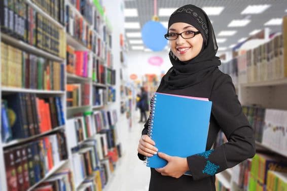 Arab student stands in the library with a notebook in hand.