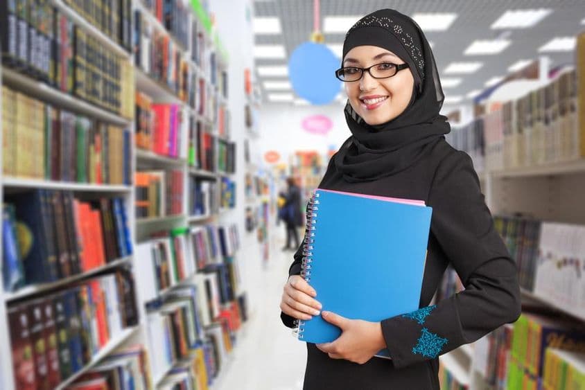 Arab student stands in the library with a notebook in hand.