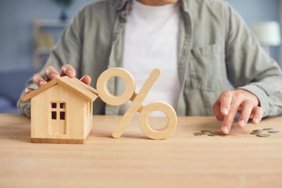 Wooden percentage symbol and house model on a table with a young man counting coins in the background.
