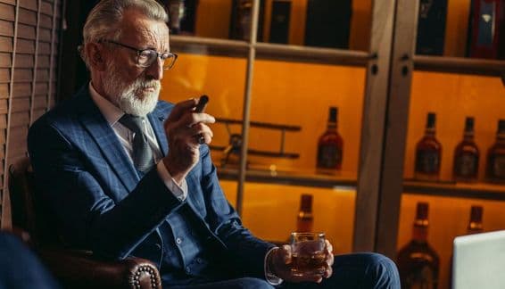 Elderly retired man using a laptop, smoking, holding expensive liquor.