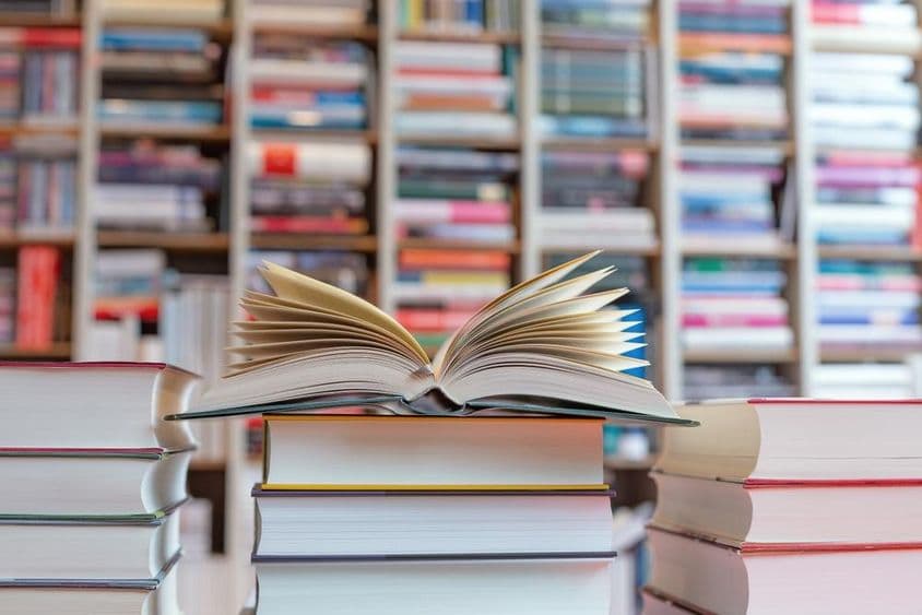 Open novel on top of a stack of books in front of a shelf.