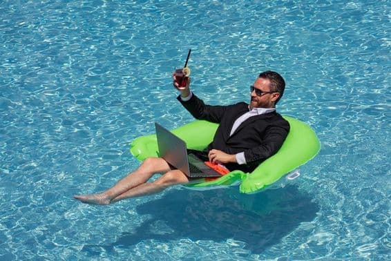 Successful businessman floating in a suit in pool water, drinking a summer cocktail and using a laptop in the pool.