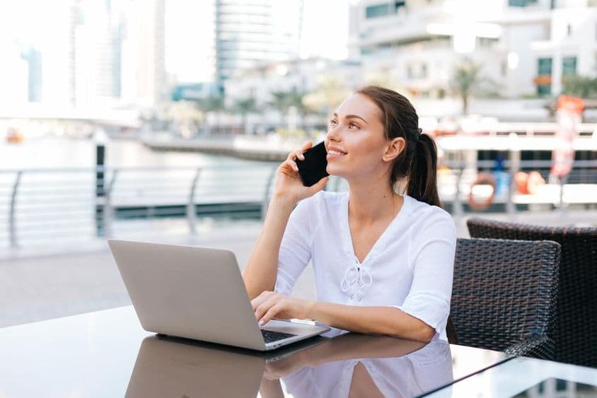 Young freelance woman working on a laptop, sitting in an outdoor cafe holding a phone.