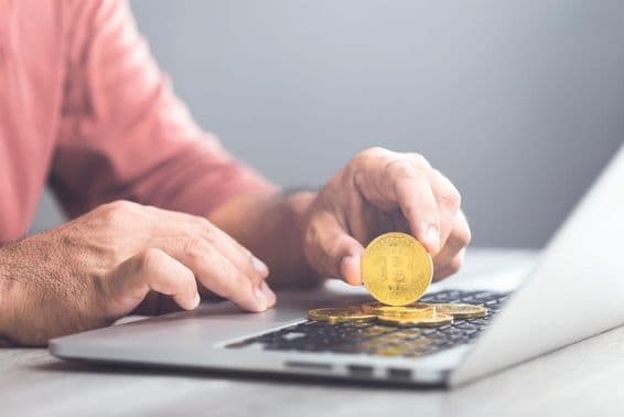 Man holding a gold-colored Bitcoin while using a laptop.