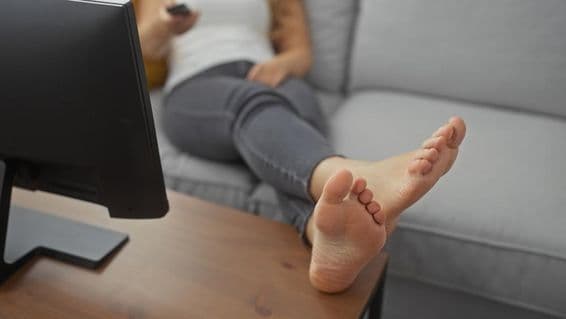 A young woman lounging barefoot on a cozy living room couch, holding a remote.