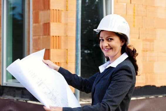 Female architect with blueprint in front of a house under construction, wearing a safety helmet.