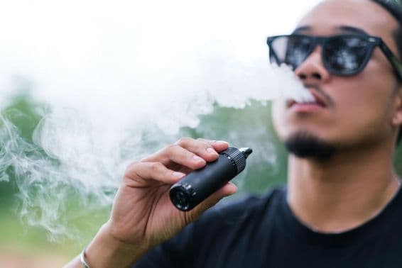 Asian man smoking with a vaping e-cigarette, exhaling smoke from his mouth and enjoying outdoors in summer.