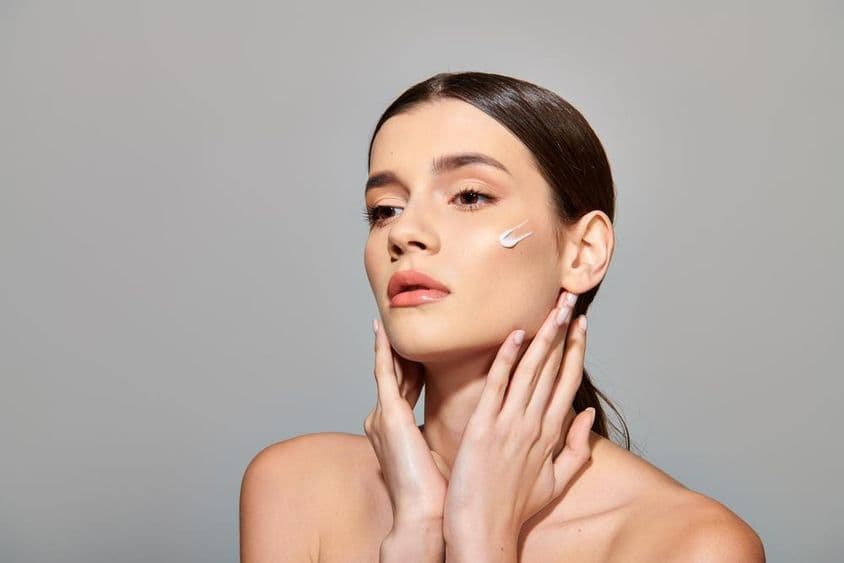 A young woman with brown hair applies a beauty cream to her face, exuding a sense of self-care.