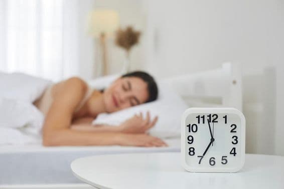 Alarm clock on bedside table in a bedroom, with a young woman sleeping in the background.