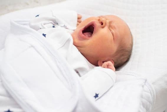 One-week-old baby boy yawning on a white bed.