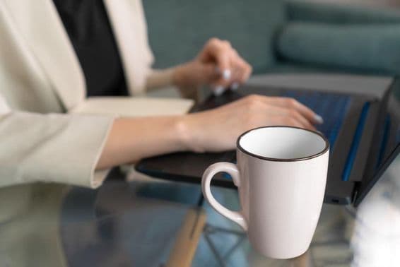 A cup on a glass table near a laptop, with a young woman typing.